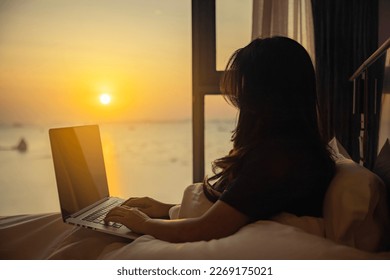 Woman sitting on bed under white blanket near window, looking to Beautiful sunset or sunrise above sea, reflection of sun in the water, surfing on internet on laptop, thoughts thinking and planning.