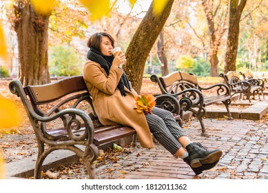 woman sitting on the bench at autumn city park drinking coffee