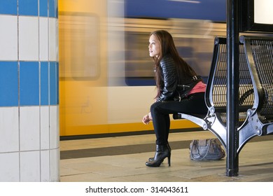 Woman, Sitting On A Bench, With Her Purse On The Platform Next To Her, Looking At A Passing Train, Waiting.