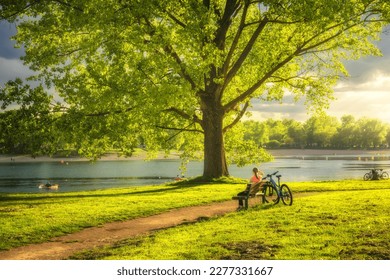 Woman sitting on bench and mountain bike, green trees and grass, lake at sunset in spring. Colorful landscape with resting girl, bicycle, river in park in summer. Sport and travel. Biking. Nature - Powered by Shutterstock