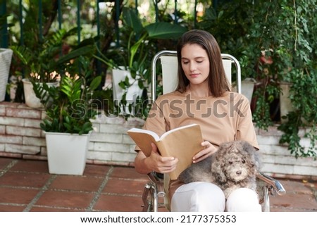 Woman sitting on chair in backyard with dog on her laps and reading book