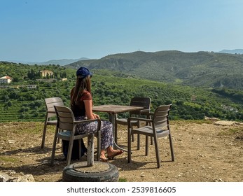 Woman sitting on chair at small table on a gravel terrace, overlooking picturesque landscape in Berat, Albania, Europe. Peaceful outdoor dining area. Rolling hills, olive groves and mountain Tomorr - Powered by Shutterstock