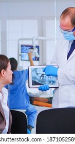 Woman Sitting On Chair In Waiting Room Of Crowded Stomatological Clinic Listening Doctor Looking On Tablet With Digital X-ray. Dentist Showing To Patient Teeth Radiography Using Modern Gadget.