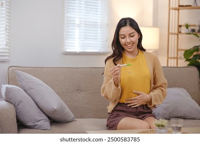 A woman is sitting on a couch holding a pregnancy test. She is smiling and she is happy about the result - Powered by Shutterstock