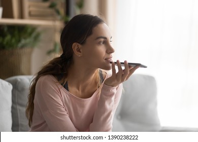 Woman sitting on couch holds phone talks on speakerphone with friend, makes voice recognition or request uses internet services through virtual assistant, record audio message, translator app concept - Powered by Shutterstock