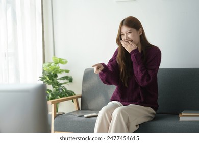 A woman sitting on a couch, laughing and pointing at a TV screen in a cozy living room with natural light and a potted plant. - Powered by Shutterstock