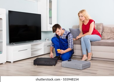 Woman Sitting On Couch Looking At Male Technician Installing TV Set Top Box At Home