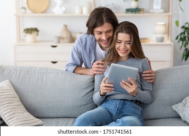 Woman sitting on couch and showing her husband content on digital tablet, home interior, copy space