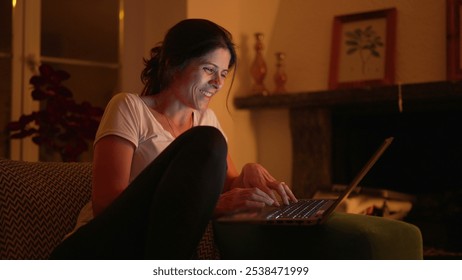 Woman sitting on a couch, smiling while working on a laptop in a dimly lit room. The warm lighting adds a cozy feel to the scene, capturing a moment of joy - Powered by Shutterstock