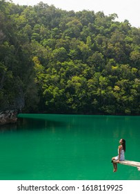 A Woman Sitting On The Edge Of A Diving Board In A Magnificent Green Lagoon.