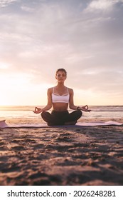 Woman sitting on exercise mat practicing yoga on the beach. Caucasian woman meditating in lotus pose.
