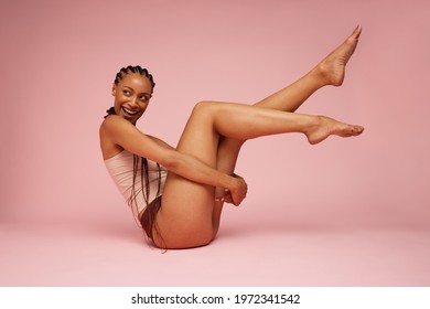 Woman sitting on floor with legs up in air. Female in underwear looking away and smiling over pink background.