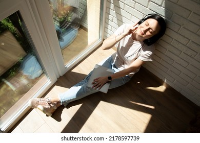 A Woman Is Sitting On The Floor Near The Window Wearing Headphones And A Laptop On Her Feet And Listening To Music With Her Eyes Closed. View From Above