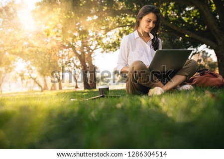 Woman sitting on grass at park working on laptop. Female wearing earphones using laptop while sitting under a tree at park with bright sunlight from behind.