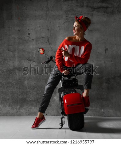 Woman sitting on motorcycle bicycle scooter  retro pinup style pointing finger at the corner in red blouse and jeans on concrete wall background looking at the corner