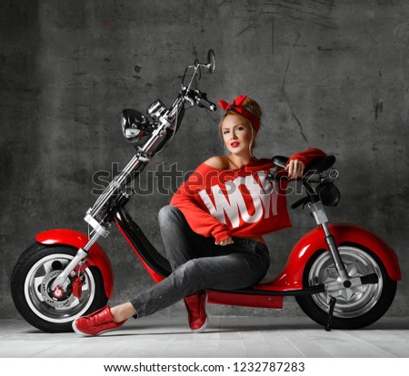 Woman sitting on motorcycle bicycle scooter  retro pinup style in red blouse and jeans on concrete wall background looking at the corner