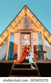 Woman is sitting on the porch of a wooden hut with lights of garlands in the evening. The concept of glamping and renting a chalet.