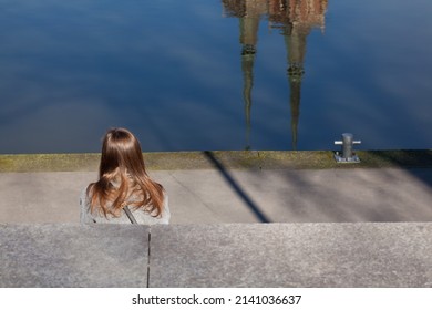 Woman Sitting On The Riverbank, City. Church Spires Reflecting On The Surface Of The Water.