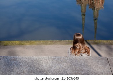 Woman Sitting On The Riverbank, City. Church Spires Reflecting On The Surface Of The Water.