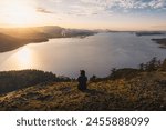 A woman is sitting on rocks and enjoying the beautiful sunset view of Mount Erskine in Salt Spring Island, British Columbia, Canada. It features a golden sky, rocks, trees, an ocean, and mountains.