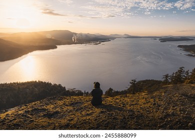 A woman is sitting on rocks and enjoying the beautiful sunset view of Mount Erskine in Salt Spring Island, British Columbia, Canada. It features a golden sky, rocks, trees, an ocean, and mountains. - Powered by Shutterstock