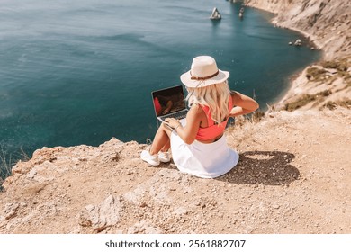 A woman is sitting on a rocky beach with a laptop open in front of her. She is wearing a pink top and a straw hat. Concept of relaxation and leisure. - Powered by Shutterstock