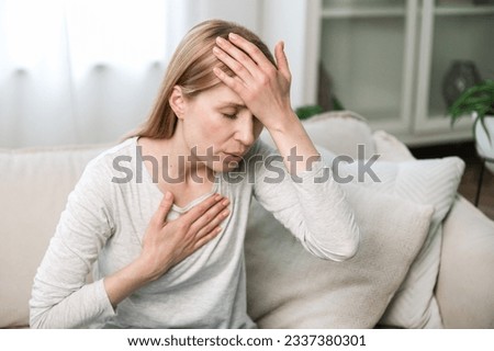 Woman sitting on sofa at home and holding hands on chest and forehead. Female having asthma, heart or panic attack. Difficulties with breathe, feeling severe pain.