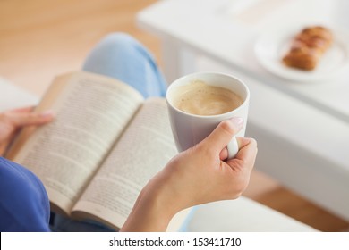 Woman sitting on the sofa reading a book holding her coffee mug in living room at home - Powered by Shutterstock