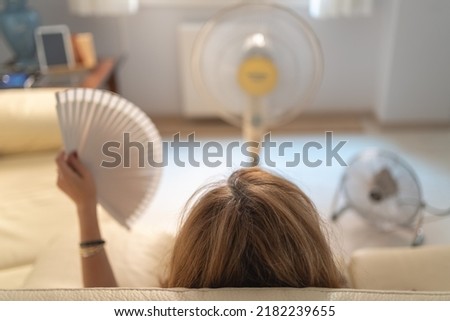 Woman sitting on a sofa with several fans giving her air and a fan in her hand, high summer temperatures.