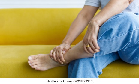 Woman Sitting On Sofa Using Hand To Hold Leg And Feeling Pain, Suffer, Hurt And Tingling. Concept Of Guillain Barre Syndrome And Numb Hands Disease Effect.