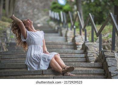 Woman Sitting On The Steps In A Long White Dress Enjoying A Summer Day. The Stairs Lead Through The Park And Are Lined With Railings. The Woman Has Slippers On Her Feet.