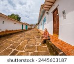 Woman sitting on a stone street in Barichara, Santander, Colombia, surrounded by traditional white colonial architecture with colorful doors and windows