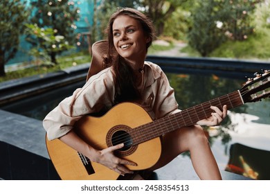 A woman is sitting on a stone wall with an acoustic guitar in front of a pond - Powered by Shutterstock