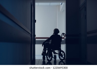 Woman Is Sitting On Wheelchair In Dark Hallway. She Is Moving In Clinic For Meeting Doctor. Copy Space In Left Side