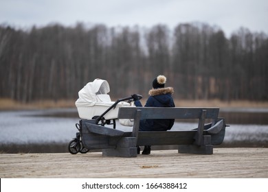 Woman sitting on wooden bench and staring at lake in cold day. White baby stroller beside young mother. Thinking about life. Back view. Peaceful atmosphere in nature.  - Powered by Shutterstock