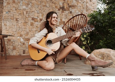 A woman is sitting on a wooden deck with an acoustic guitar and a hat on - Powered by Shutterstock