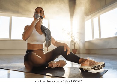 Woman sitting on yoga mat in gym, drinking water, sunlit room, relaxation - Powered by Shutterstock