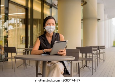 Woman Sitting Outdoors At Coffee Shop Restaurant Social Distancing And Wearing Face Mask To Protect From Covid 19 While Using Phone And Digital Tablet Horizontal Shot.