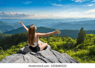 Woman sitting with outstretched arms in summer mountain scenery. Woman on hiking trip relaxing looking at beautiful mountain view. Blue Ridge Parkway, Near Asheville, North Carolina, USA. - Shutterstock ID 2258684223