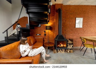 Woman sitting relaxed on a couch and using phone at home. Wide interior view in loft style with burning fireplace and dog on the stairs