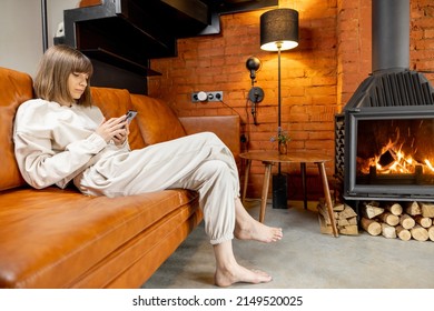 Woman Sitting Relaxed On A Couch And Using Phone At Cozy Home Interior In Loft Style With Burning Fireplace On Background