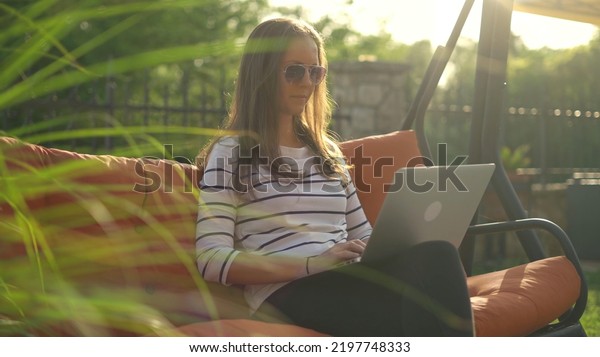 Woman sitting in swinging couch outdoor in garden\
working with laptop computer. Home office, working on vacation, sun\
light, lens flare.