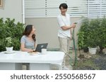 a woman sitting at a table with a laptop and a cup of coffee. The man is holding a hose and spraying water..