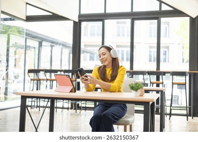 A woman is sitting at a table with a pink tablet and a cell phone. She is wearing headphones and she is listening to music. The scene suggests a relaxed and leisurely atmosphere