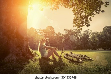 Woman Sitting Under Sun Light At Day Near Her Bicycle In The Park