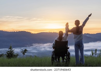 Woman sitting in wheelchair with his care helper raised hands outdoor morning sunrise mountain background. Rear view - Powered by Shutterstock