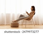 A woman is sitting in a woven chair and looking at her phone. Her feet are resting on an ottoman.