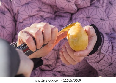 A Woman In A Ski Jacket Is Peeling A Potato With A Pocket Knife Outdoors On A Sunny Day.