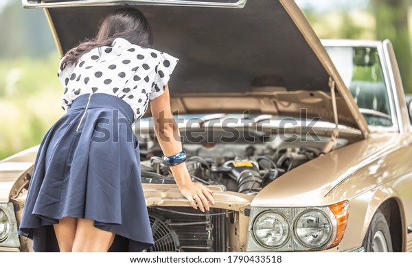 Woman in skirt leans to an open hood\
of a vintage convertible car engine after a breakdown.\

