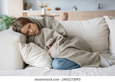 Woman sleeping on couch at home resting wrapping in plaid feeling bad symptoms. Girl suffering from seasonal flu or cold. Feel unhealthy with influenza. Tired exhausted workaholic becoming ill, sick. - Powered by Shutterstock